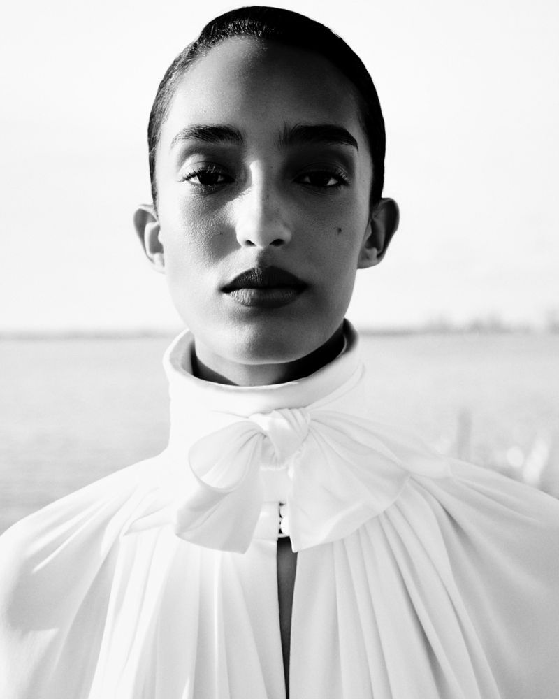 Close-up portrait of an African American woman in a white high-necked blouse with a bow, photographed outdoors against a blurred background.