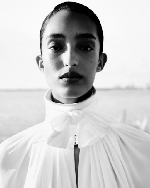 Close-up portrait of an African American woman in a white high-necked blouse with a bow, photographed outdoors against a blurred background.