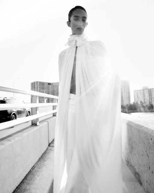 Person in flowing white outfit poses on a bridge against a cityscape, with a car visible in the background, black and white.