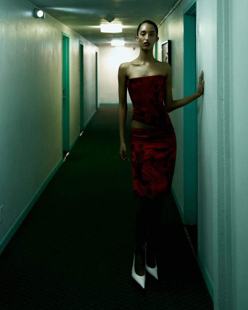 Woman in red strapless dress and white heels poses in a hallway with teal doors and green carpet, looking elegant and fashionable.