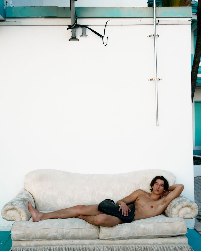 Man lounges shirtless on a white vintage sofa outdoors, leaning against a white wall with overhead pipes and a view of palm trees.