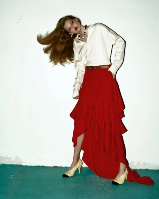 Fashion editorial shot of a young woman posing in a white blouse and red tiered skirt, set against a white wall and teal floor.
