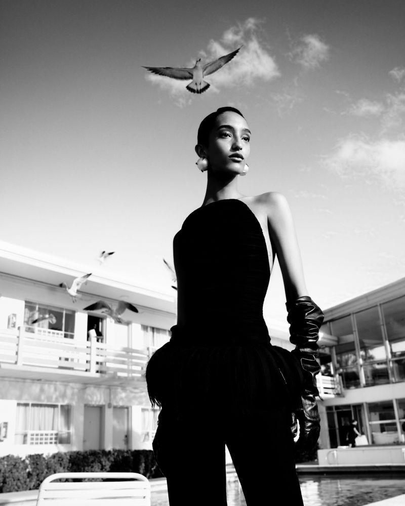 Black and white fashion shot of a woman in a dark strapless dress, gloves, and earrings, with a motel and birds in the background.
