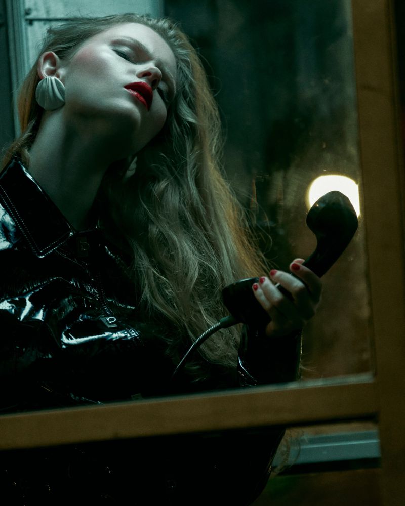 Fashionable woman with blonde wavy hair, red lipstick, and a black patent-leather jacket, holding a vintage black telephone receiver indoors.
