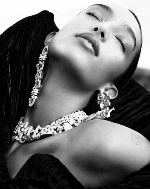Black and white close-up of a woman with closed eyes, wearing dramatic jewelry, including earrings and a large necklace, in a studio.