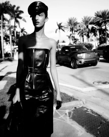 Model wearing a leather cap, strapless top, and skirt standing outdoors on a sunny street with palm trees and a parked truck in the background.