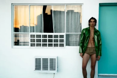 A man wearing an open green jacket leans against a white wall next to a teal door and large window.
