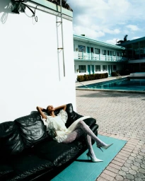 Model reclining on a black leather sofa outdoors next to a motel pool, wearing a white ruffled dress and patterned tights.