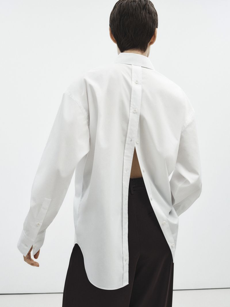 Person from the back wearing an oversized white button-down shirt with a brown pair of pants against a neutral background.