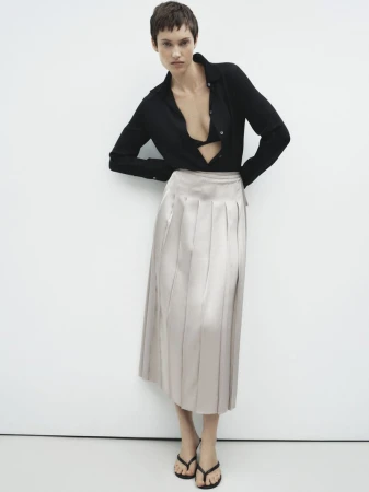 Model with short dark hair wearing a black blouse and pleated cream skirt against a plain white background