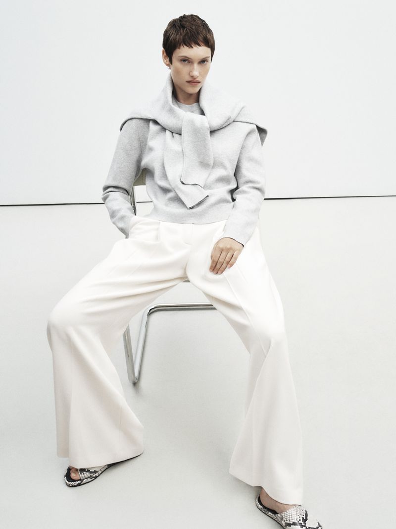 Adult woman in grey sweater and white slacks sitting in studio with a sweater draped over her shoulders, fashion editorial.