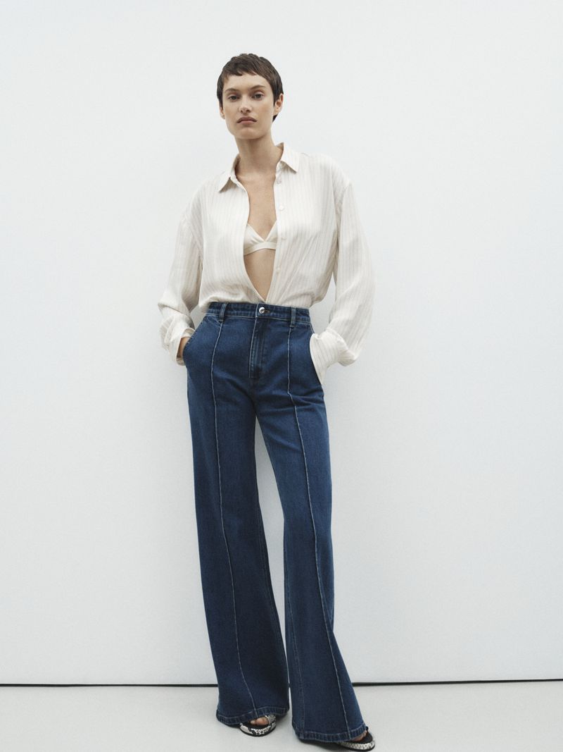 A woman with short hair poses in a beige blouse and wide-leg jeans against a white background in a studio shot, showcasing fashion apparel.