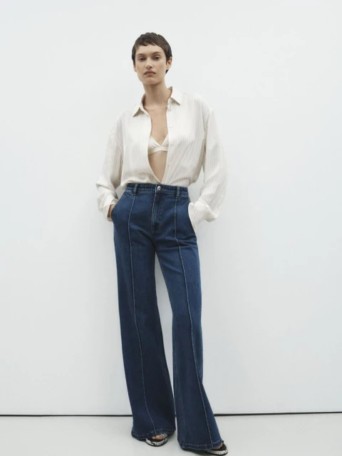 A woman with short hair poses in a beige blouse and wide-leg jeans against a white background in a studio shot, showcasing fashion apparel.