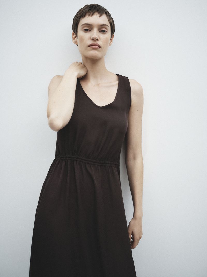 Model with short dark hair wearing a sleeveless dark brown dress against a plain white wall