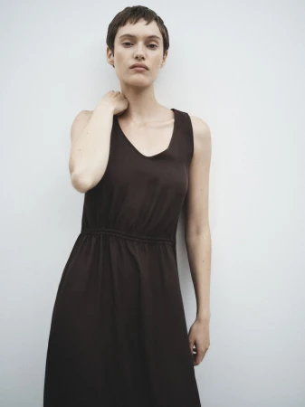 Model with short dark hair wearing a sleeveless dark brown dress against a plain white wall