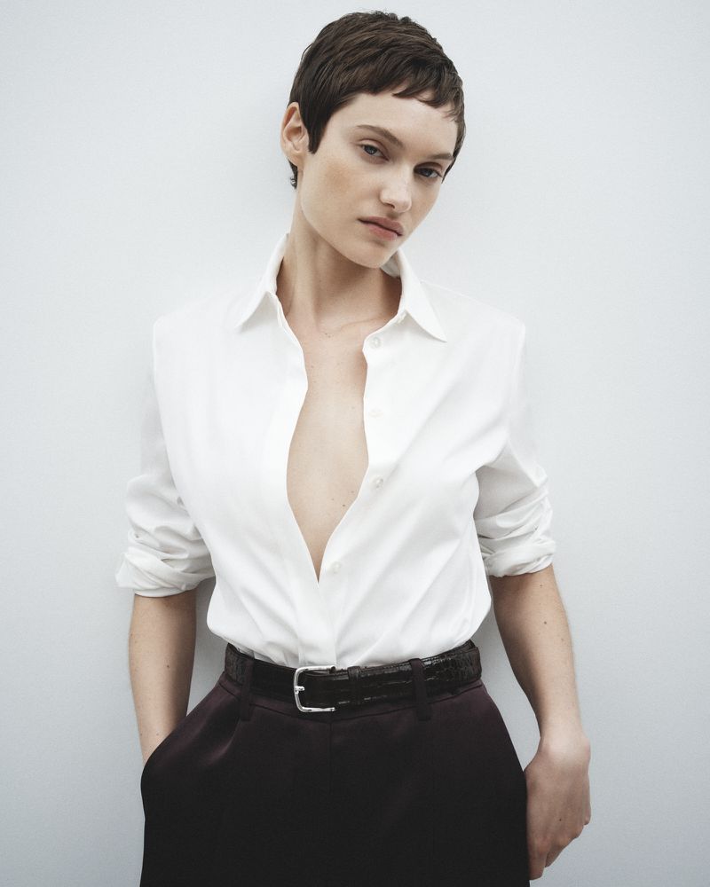Elegant woman in a white blouse and dark trousers, posing against a white backdrop in a fashion-forward studio portrait.