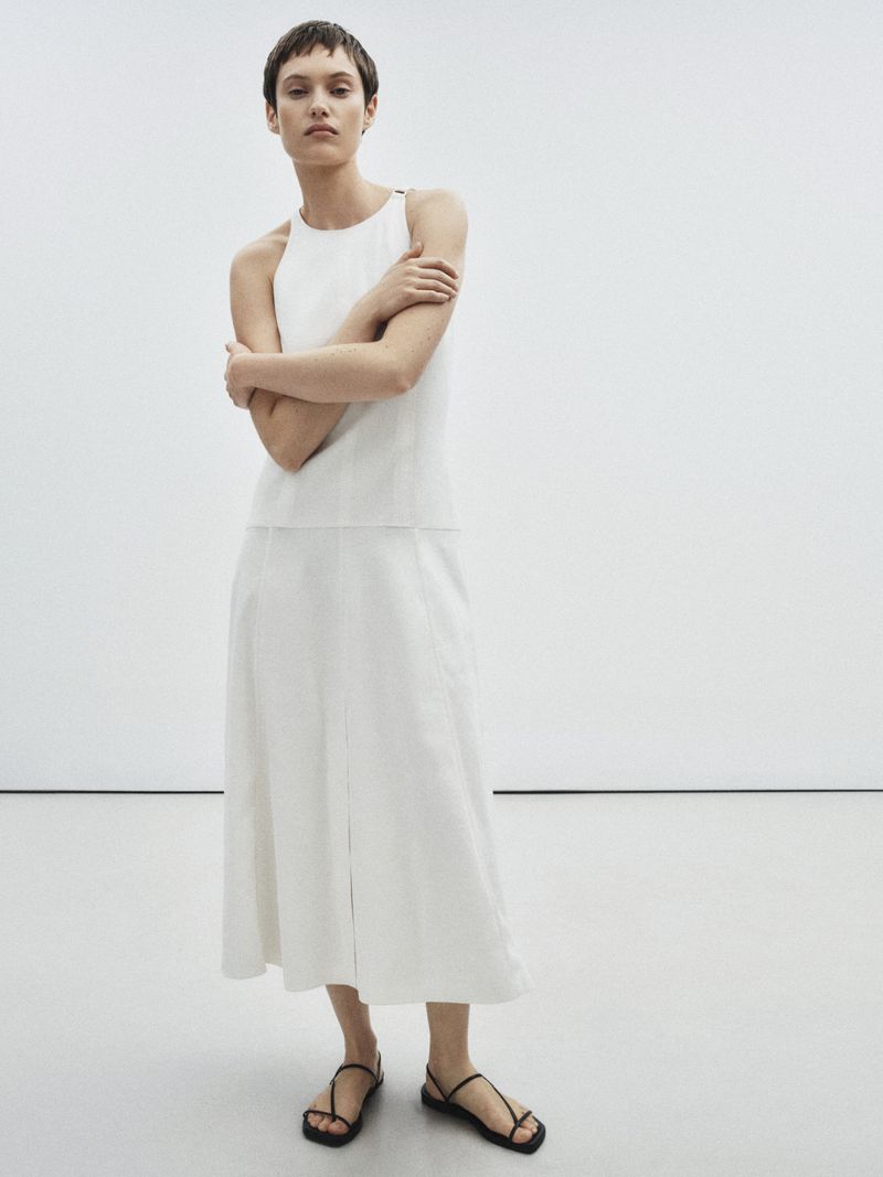 Woman in a white top and skirt poses in front of a white wall. She has short hair, crossed arms, and is wearing black sandals.