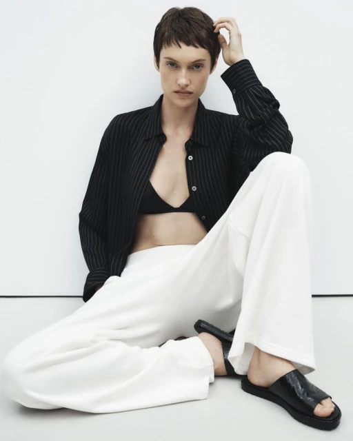 Fashion model wearing a striped button-down shirt and white pants sitting against a white wall in a studio environment.