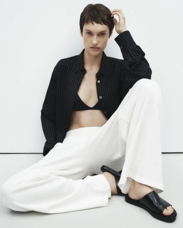 Fashion model wearing a striped button-down shirt and white pants sitting against a white wall in a studio environment.