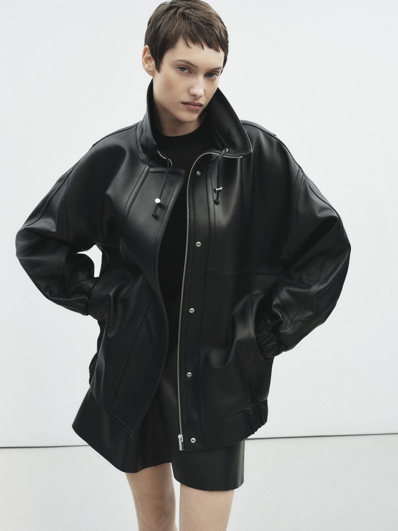 A young adult woman poses in a studio, wearing a black, oversized leather jacket and shorts, showcasing fashion and editorial style.