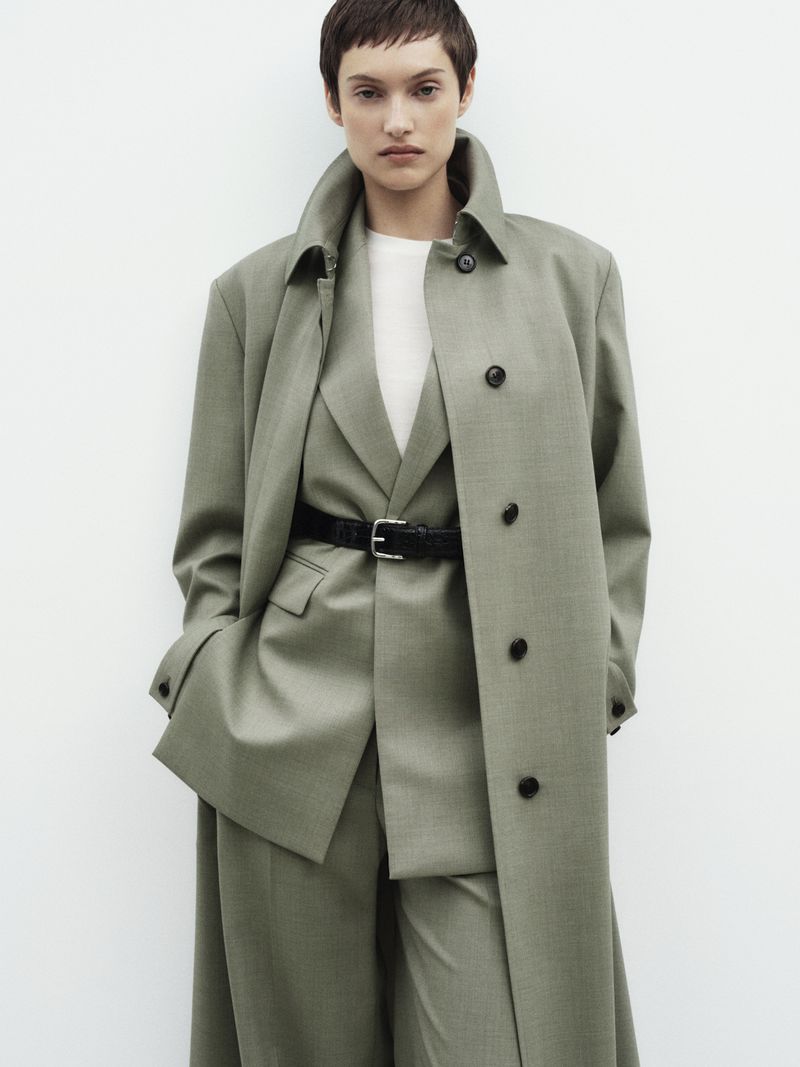 Woman in olive green trench coat, suit jacket, pants, and black belt against a white background. Fashion editorial style.
