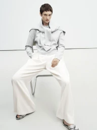 Model seated on a chair wearing a grey sweater with another tied over shoulders and white trousers against a white wall.