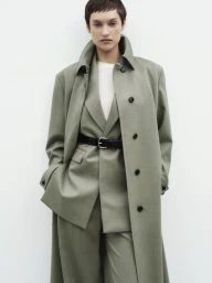 Model with short dark hair wearing a sage green coat over a matching suit against a plain white background