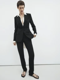 Model with short dark hair wearing a black pantsuit and white collared shirt against a white studio background.