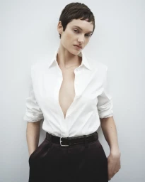Model with short dark hair wearing a white collared shirt and dark trousers against a plain light background