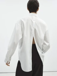 Model seen from behind wearing an oversized white button-down shirt with a buttoned placket running down the back over dark trousers.