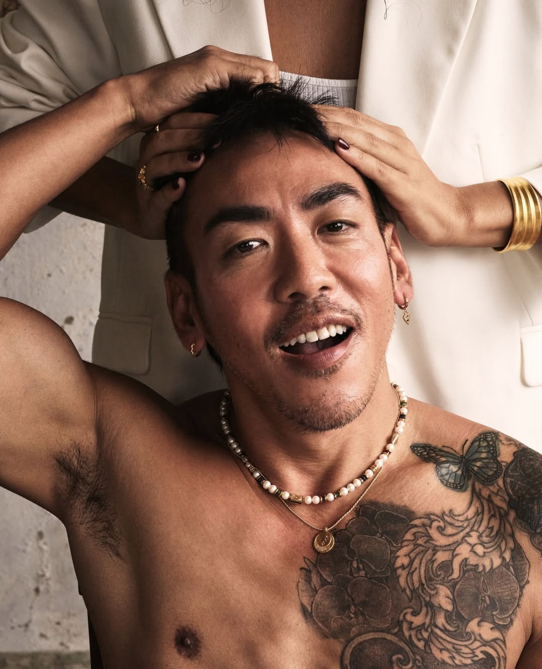 Close-up of an adult Asian male with a tattoo and jewelry. Someone is adjusting his hair. The background features a white wall and jacket.