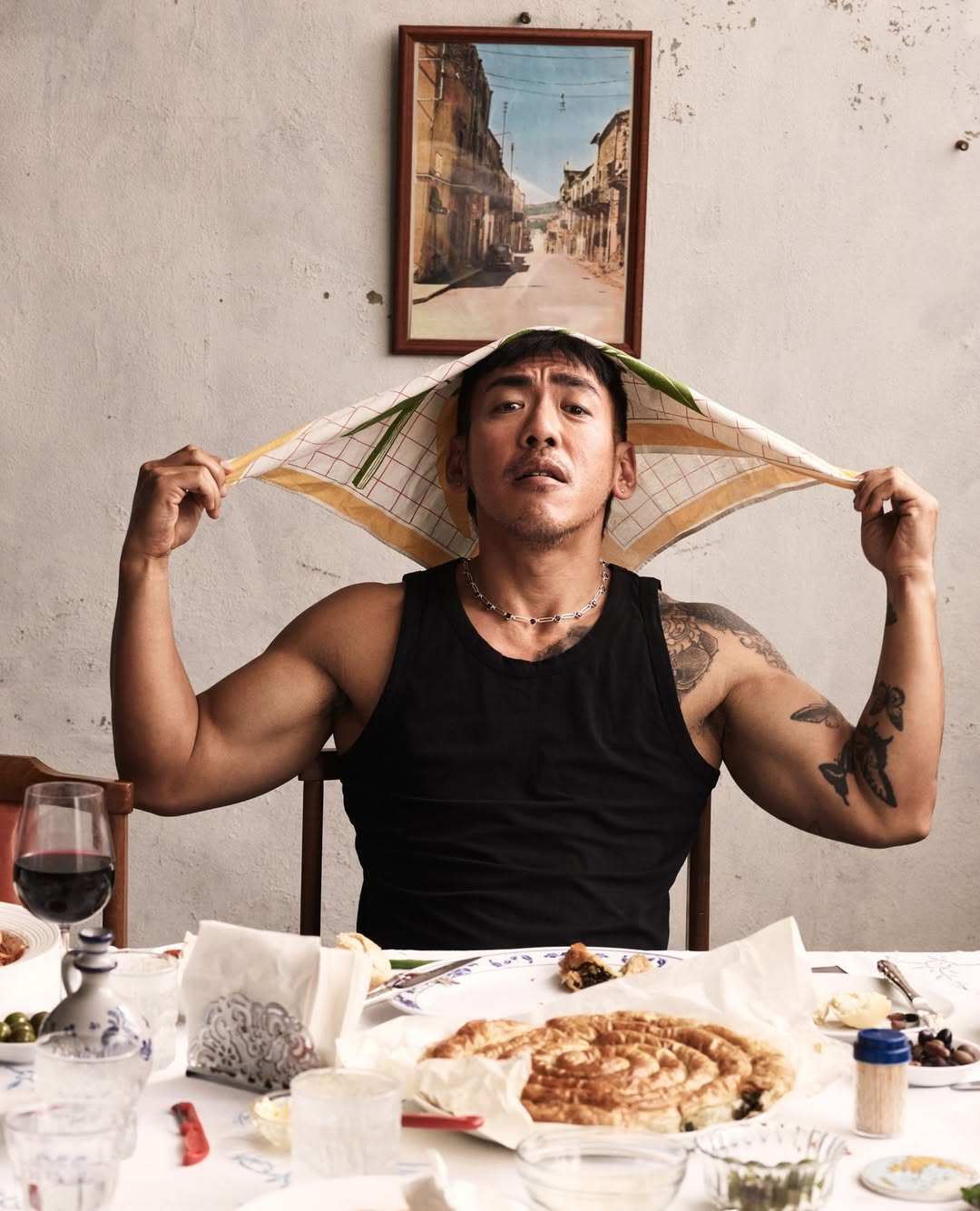 Adult male indoors with a towel on his head, sitting at a table with food and a glass of wine, and a landscape painting on the wall.