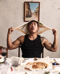 Man wearing a black tank top seated at a dining table holding a patterned cloth over his head like a hat