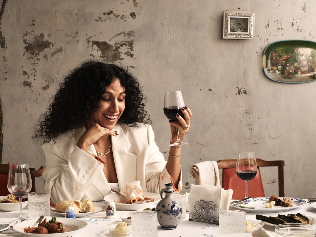 Woman with curly hair enjoys a glass of red wine at a table laden with food and drinks, set against a textured wall backdrop.