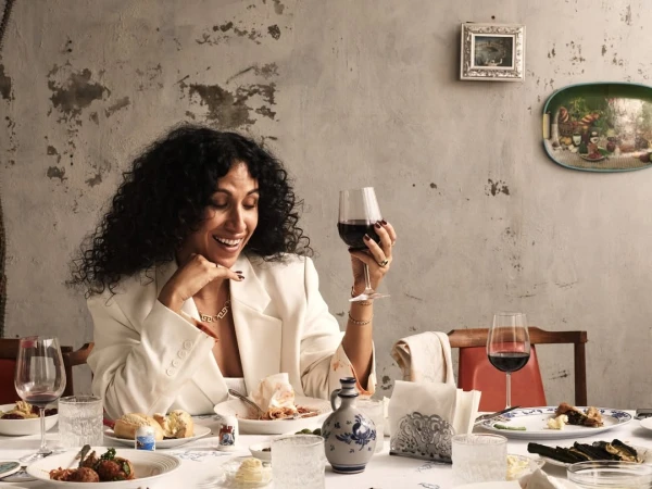 Woman with curly hair enjoys a glass of red wine at a table laden with food and drinks, set against a textured wall backdrop.