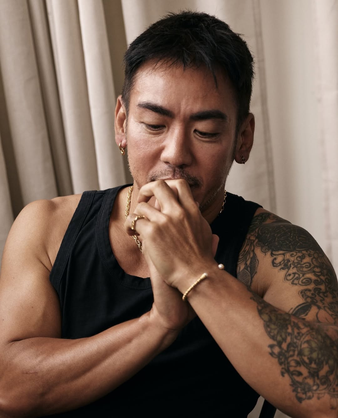 Portrait of an adult male with tattoo on arm, wearing a black tank top and gold jewelry, sitting indoors with neutral background.