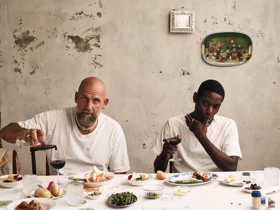 Two men, one bald with a beard and one dark-skinned, are seated at a table filled with food and wine in a rustic indoor setting.