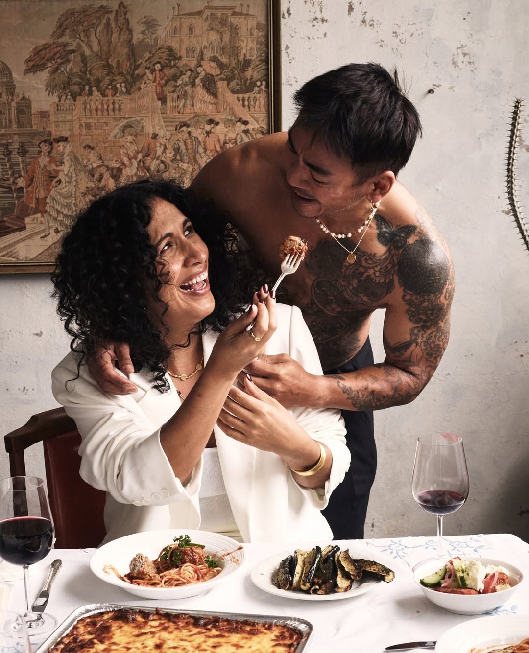 Woman in a white blazer feeding an Asian man a meatball on a fork at a dinner table with various dishes, glasses of red wine, and flatware.