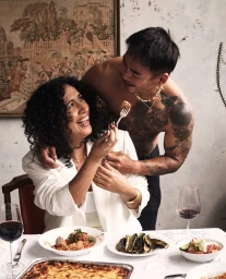 A smiling woman in a white blazer and a shirtless tattooed man sharing food over a dinner table laden with Italian dishes.