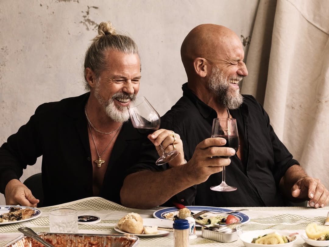 Two adult men with beards and black shirts are seated at a table with food and wine, laughing and enjoying each other's company in an indoor setting.