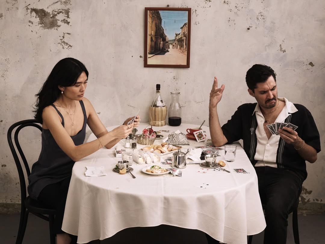 Two people, an Asian woman and a Caucasian man, sit at a table covered with a white tablecloth, playing cards in an indoor setting.