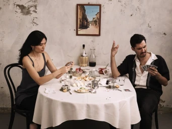 A man and woman sitting at a round dining table playing cards against a rustic distressed wall for Temple of the Sun.