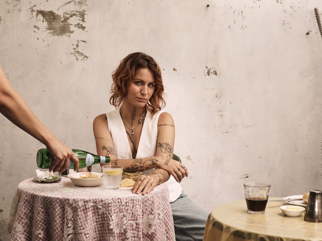 Woman sits at a table with food and a water glass, with a person pouring a bottle of water in the background; rustic indoor setting.