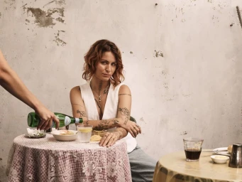 Woman in white sleeveless top with tattoos seated at a table as a hand pours water from a green bottle