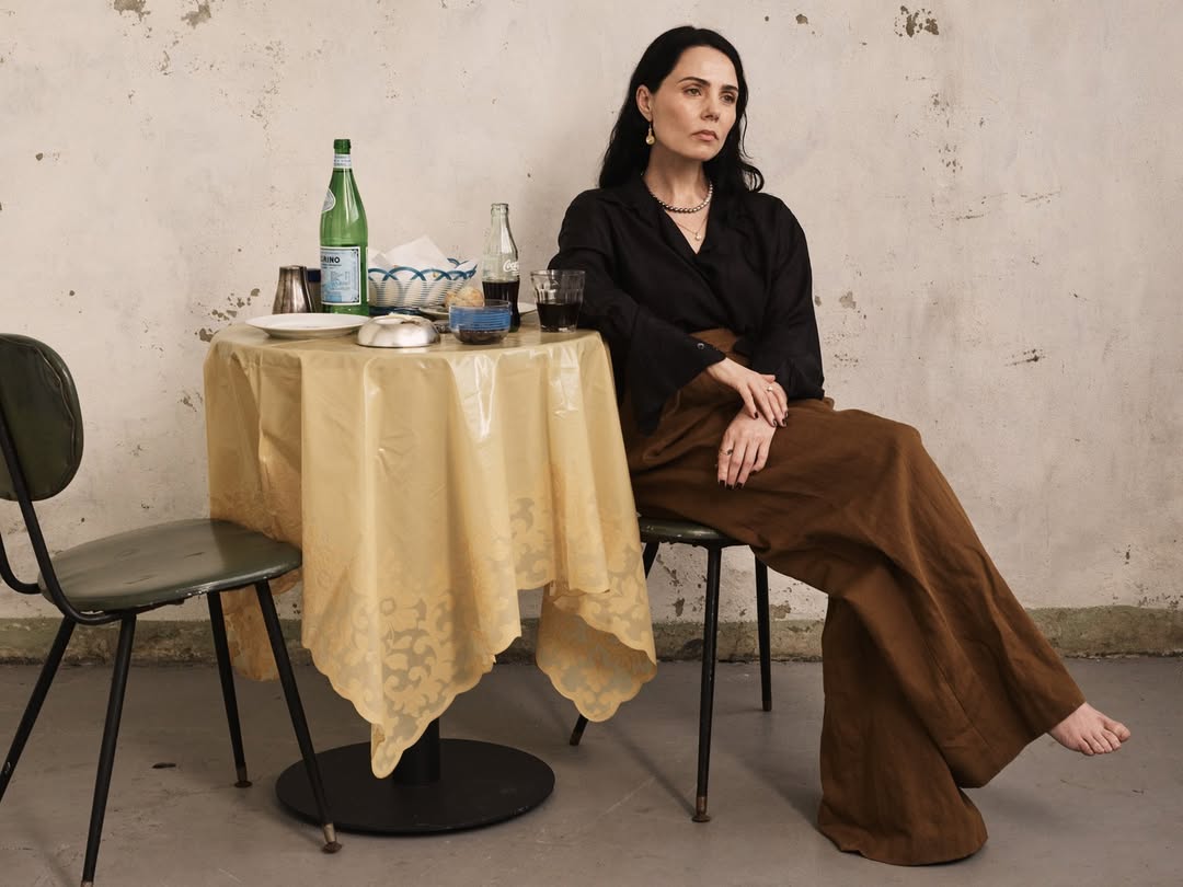 Adult woman with dark hair and a serious expression sits at a table in an indoor setting in a medium shot with natural lighting.