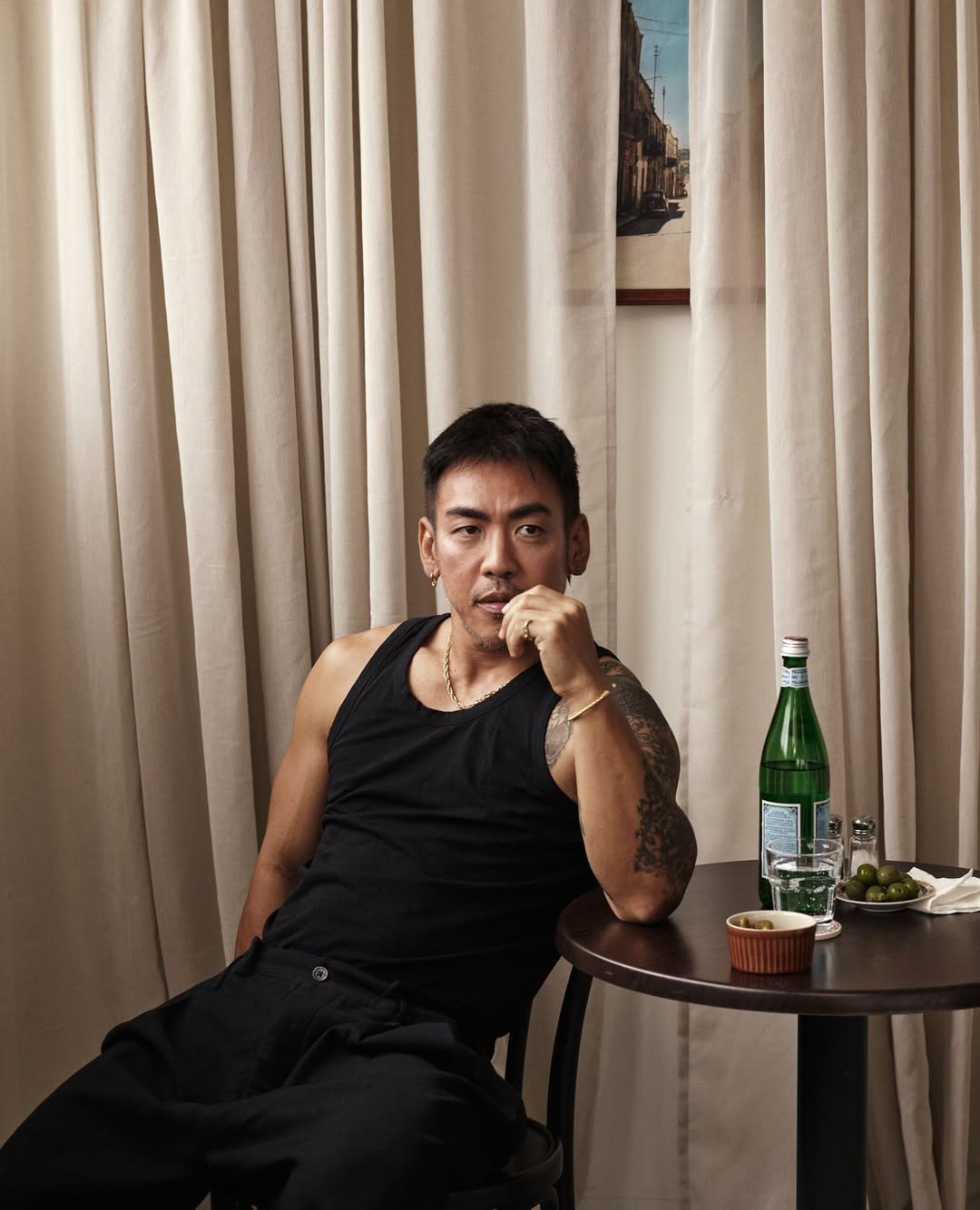 Portrait of a tattooed adult male in a black tank top sitting at a table indoors with a bottle of sparkling water and various dishes.