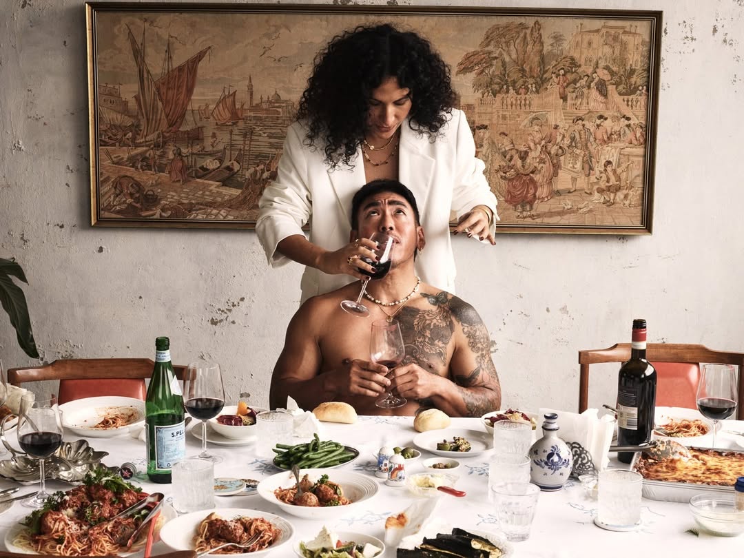 Two people at a dining table with food and drinks, featuring a woman in a white blazer pouring red wine for a man.