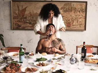 Woman in white jacket standing behind a shirtless man seated at a banquet table filled with food and wine for Temple of the Sun