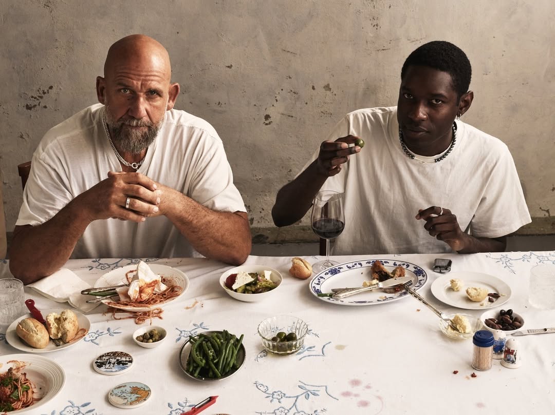 Two men at a table covered with dishes of food, including salad, bread, and green beans, with a rustic wall in the background.