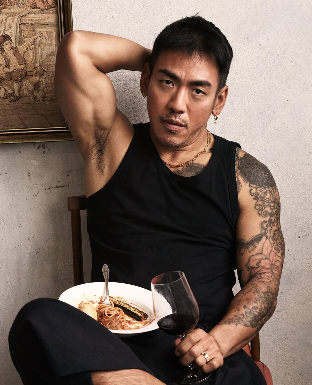 Muscular adult male with tattoos sits indoors wearing a tank top, holding a plate of spaghetti and a wine glass; an art-filled background.